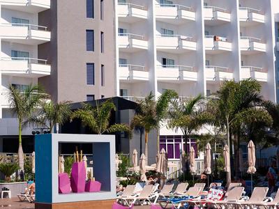 Hotel with pool, palm trees, and sun umbrellas in front of a multi-story building under a blue sky.