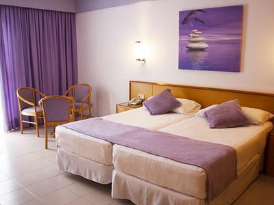 Jasny pokój hotelowy z dwoma łóżkami pojedynczymi, fioletowymi poduszkami i zasłonami oraz miejscem do siedzenia.