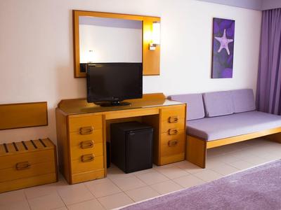 Pokój hotelowy z telewizorem, sofą, lustrem, stolikiem nocnym i fioletowymi zasłonami.