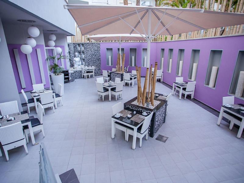 Nowoczesny zewnętrzny obszar hotelowy z fioletowymi ścianami, białymi stołami i krzesłami pod dużymi parasolami.