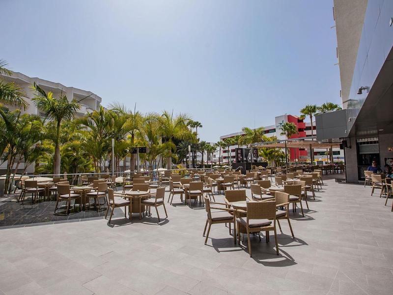 Duży taras na zewnątrz z wieloma stołami i krzesłami, otoczony palmami pod czystym niebem.