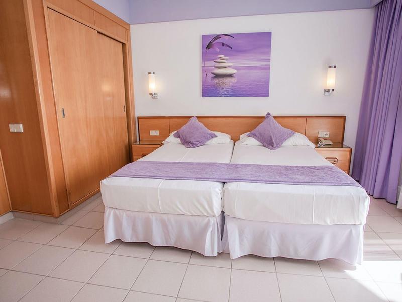 Podwójne łóżko z fioletowymi poduszkami i zasłonami w jasno urządzonym pokoju hotelowym.