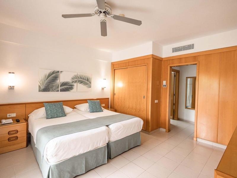 Jasny pokój hotelowy z dwoma łóżkami, drewnianymi meblami i wentylatorem sufitowym.