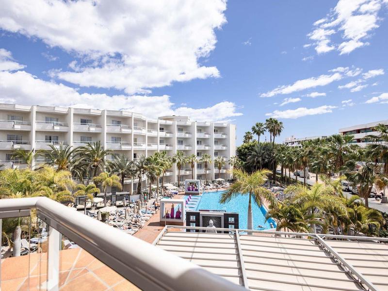 Hotel z dużym basenem otoczonym palmami i białymi budynkami pod niebieskim niebem.