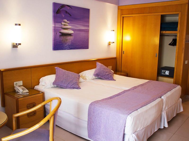 Przytulny pokój hotelowy z podwójnym łóżkiem, fioletowymi poduszkami, drewnianymi meblami i otwartą szafą.