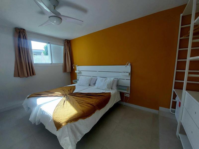 Jednoduchý hotelový pokoj s manželskou postelí, oranžovou zdí a oknem se závěsy.