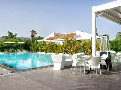 Area esterna moderna con piscina, sedie e tavoli bianchi, circondata da palme e edifici.