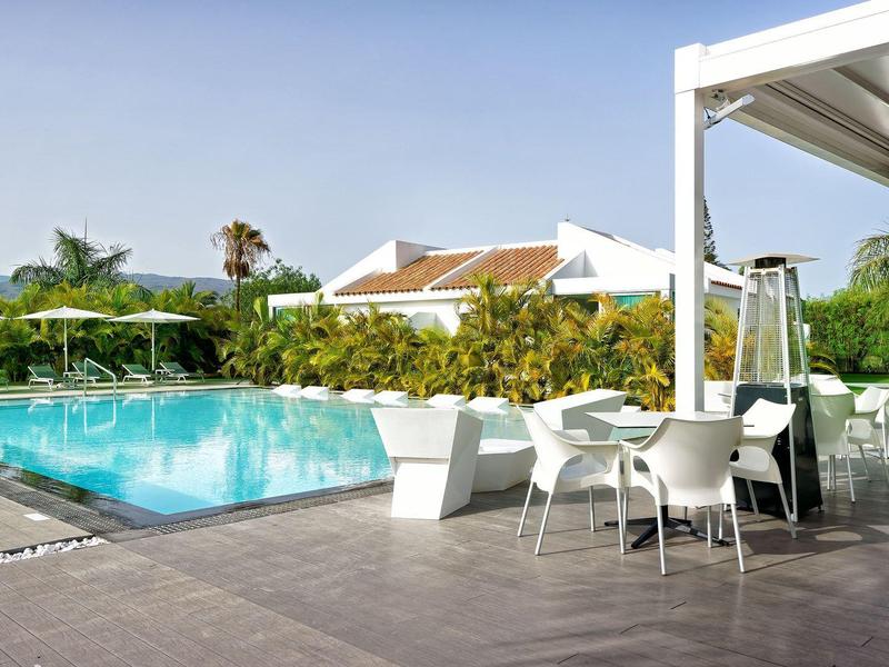 Moderní venkovní prostor s bazénem, bílými židlemi a stoly obklopený palmami a budovami.