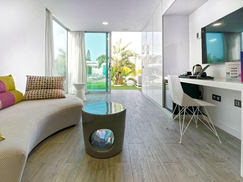 Světý hotelový pokoj s moderní pohovkou, pracovním stolem a skleněnými dveřmi na balkon s výhledem na bazén.