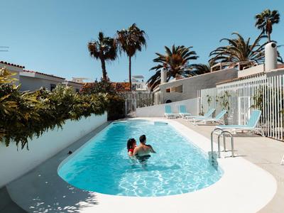 Zwei Personen schwimmen in einem Außenpool mit Palmen und weißer Terrasse bei klarem Himmel.