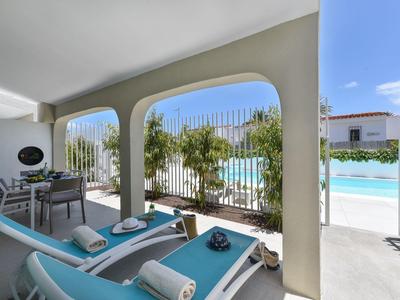 Overdekt terras met twee blauwe ligstoelen en uitzicht op het zwembad bij een heldere hemel.