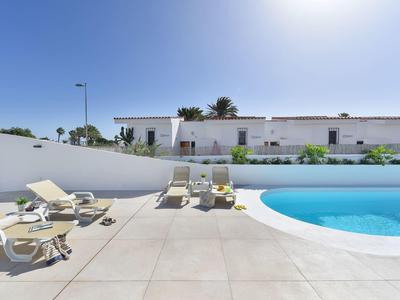 Modern buitenzwembad met ligstoelen onder een heldere blauwe lucht en wit gebouw op de achtergrond.