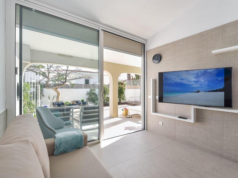 Moderne woonkamer met bank, aan de muur gemonteerde tv en toegang tot terras met tuinmeubilair.