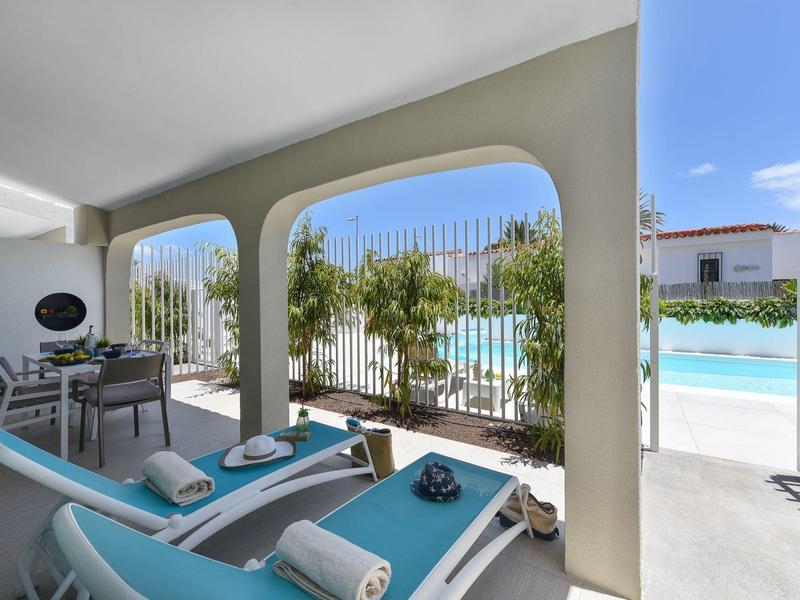 Overdekt terras met twee blauwe ligstoelen en uitzicht op het zwembad bij een heldere hemel.