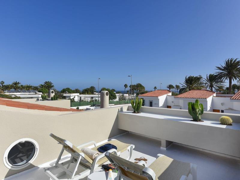 Dakterras met ligstoelen, uitzicht op witte huizen en palmbomen onder een blauwe lucht.