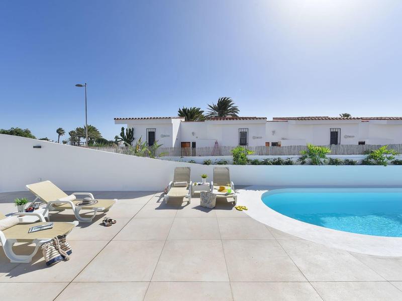 Modern buitenzwembad met ligstoelen onder een heldere blauwe lucht en wit gebouw op de achtergrond.