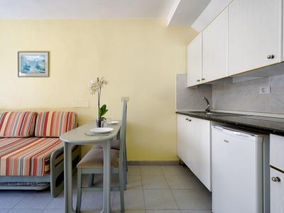 Kleine woonkeuken met slaapbank, eettafel en witte keukenkastjes in een hotelkamer.