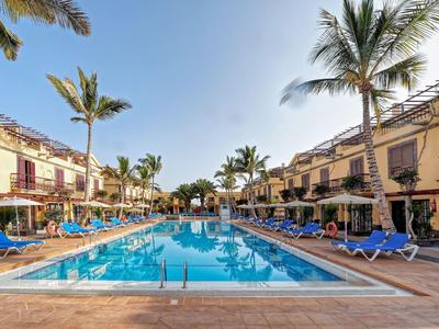 Een groot buitenzwembad omringd door ligstoelen en palmbomen, geflankeerd door hotelgebouwen van twee verdiepingen.
