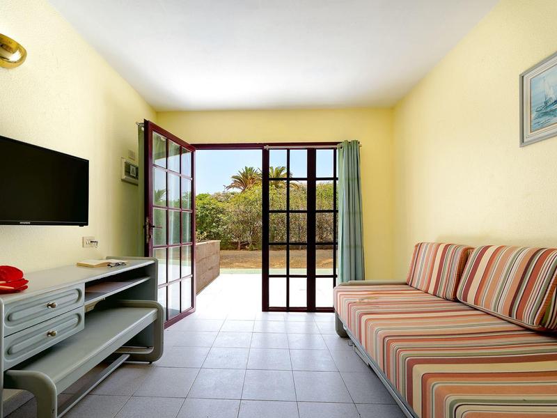 Lichte woonkamer met bank, tv en open deur naar balkon met uitzicht op bomen.