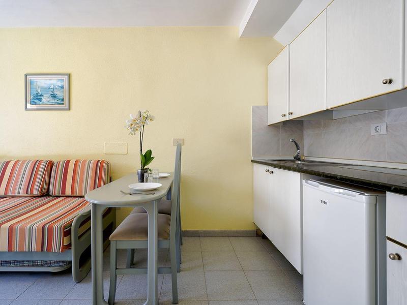 Kleine woonkeuken met slaapbank, eettafel en witte keukenkastjes in een hotelkamer.