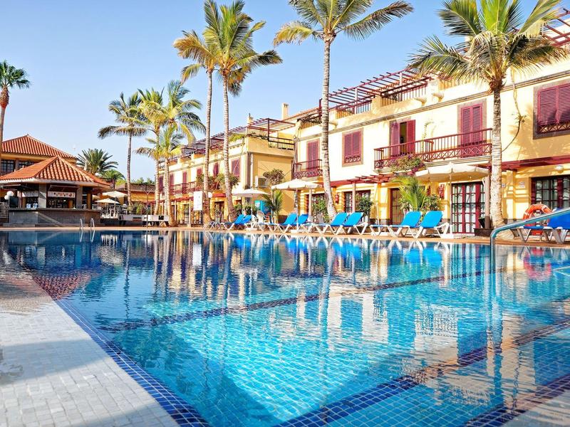 Hotel met groot zwembad, blauwe ligstoelen en palmbomen onder een heldere hemel.