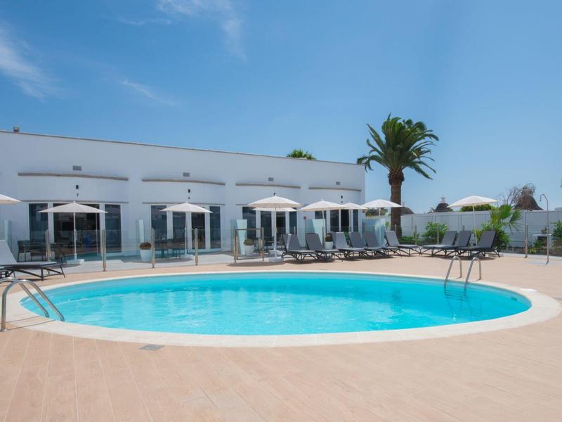 Kulový bazén s lehátky a slunečníky před bílou budovou pod slunečnou oblohou.