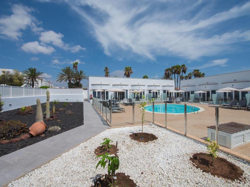 Moderní hotelový bazén s bílými budovami, palmami a modrou oblohou.