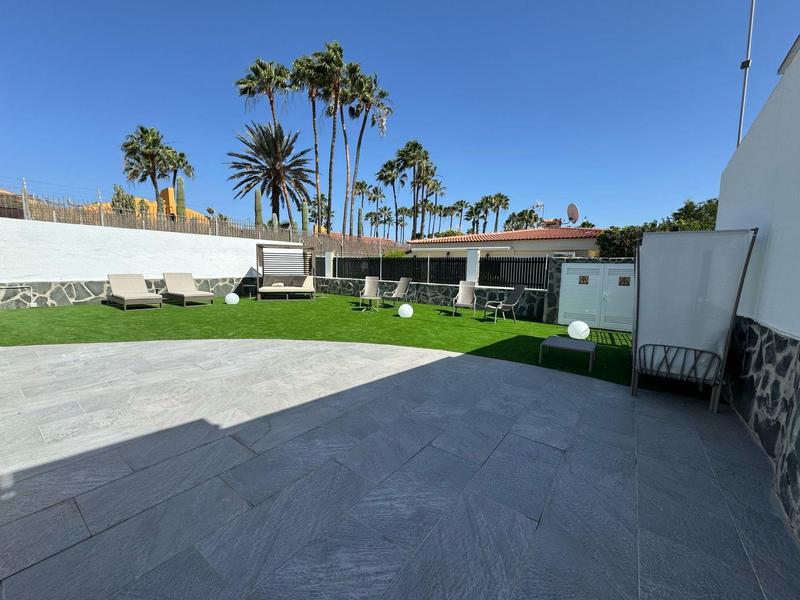 Velká hotelová terasa s šedou dlažbou, trávníkem a palmami pod modrou oblohou.