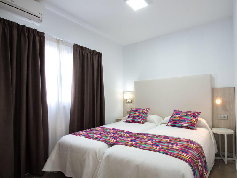 Moderně zařízený hotelový pokoj s manželskou postelí, barevnými polštáři a tmavými závěsy.