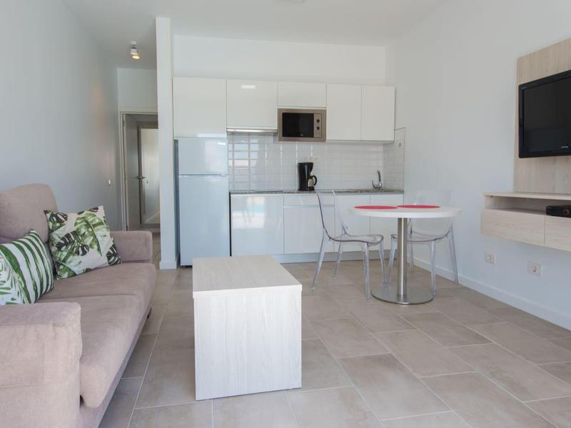 Moderně zařízený obývací pokoj s béžovou pohovkou, kuchyní a televizí na stěně.