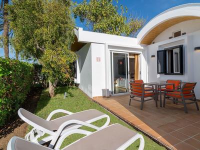 Weißes Haus mit Terrasse, zwei Stühlen, Tisch und Liegestühlen, umgeben von grünem Garten.