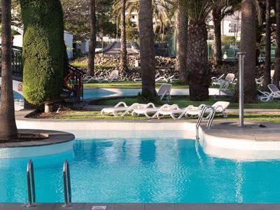 Area piscina limpida con lettini e alberi sullo sfondo di un hotel.