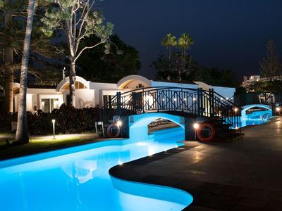 Piscina illuminata con un piccolo ponte e alberi di notte in un giardino di un hotel.