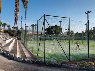 Freiluft-Tenniscourt mit grünem Belag, Umzäunung und einer Person im hellen Outfit.