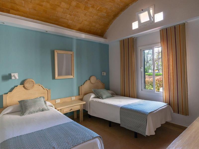 Zweibettzimmer mit blauen Wänden, Holzmöbeln, großen Fenstern und vorhängen, warmes Tageslicht.