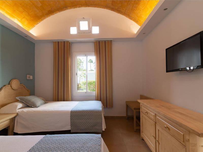 Zimmer mit zwei Einzelbetten, Holzmöbeln, Fenster mit Vorhängen und Deckenlampe unter gewölbter Decke.