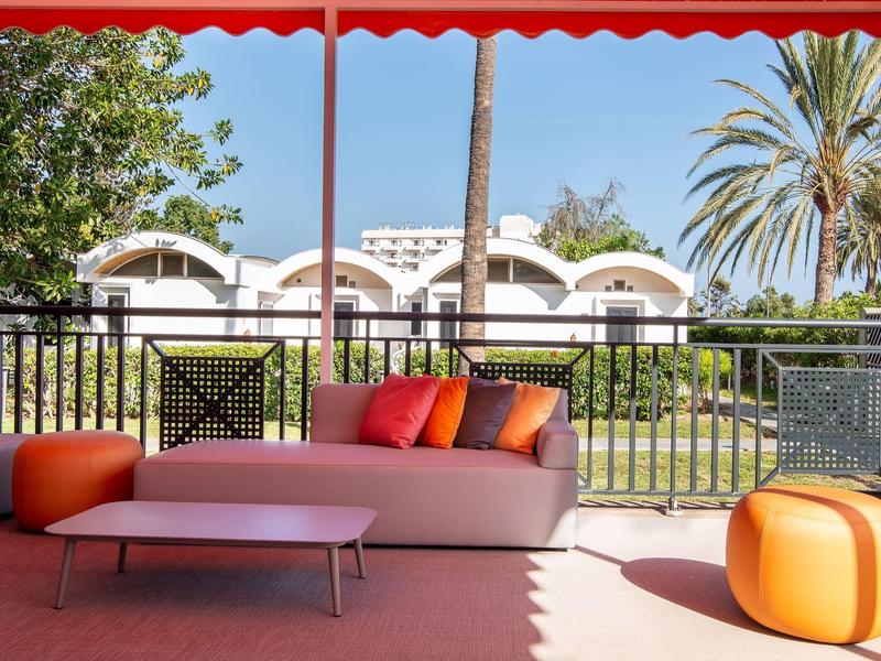 Moderner Außenbereich mit gemütlicher Couch, bunten Kissen, Orchidee und Palmen im Hintergrund.