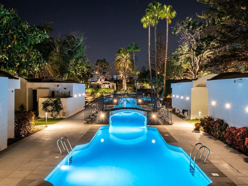 Beleuchteter Pool mit Wasserfontäne und Palmen in einer modernen, ruhigen Gartenanlage bei Nacht.