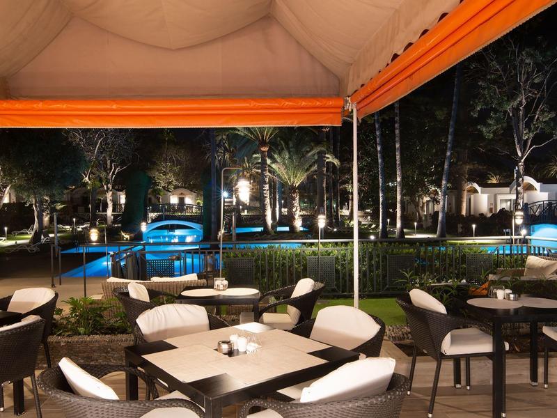 Terrasse mit Tischen und Stühlen, beleuchteter Pool und Garten bei Nacht, warme Lichter.