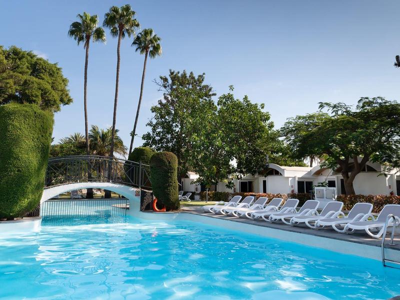 Schwimmbad mit klarem blauen Wasser, Liegestühlen, Palmen und grünem Pflanzenbereich im Sonnenschein.