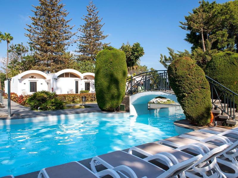 Schwimmbad mit blauem Wasser, weißen Liegestühlen, grünen Sträuchern und weißem Gebäude im Hintergrund.