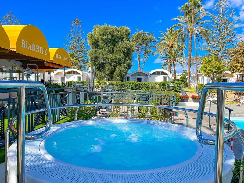 Runder Whirlpool mit blauem Wasser, umgeben von Metallgeländern und grüner Gartenlandschaft.