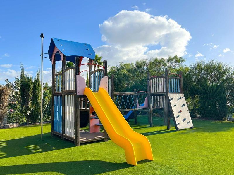 Bunter Spielplatz mit gelber Rutsche, Kletterwand und grünem Kunstrasen bei blauem Himmel.