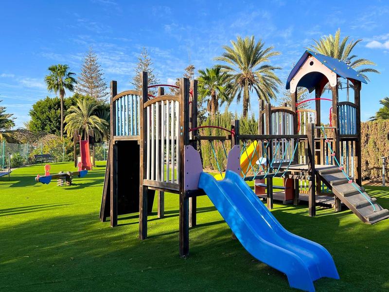Kinder-Spielplatz mit Rutsche, Klettergerüst und grüner Wiese unter blauem Himmel.