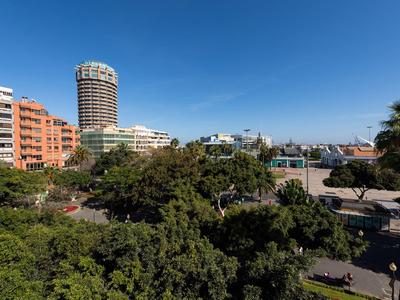 Stadtansicht mit runden Hochhaus, roten Gebäuden, vielen grünen Bäumen und klarem blauem Himmel.