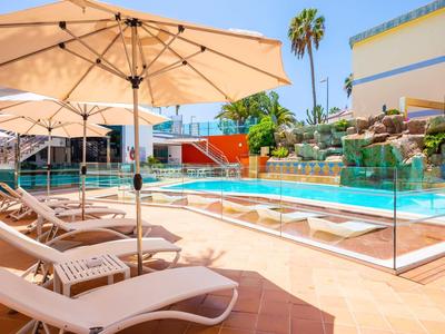 Poolbereich mit Liegestühlen und Sonnenschirmen neben einem Hotelgebäude unter blauem Himmel