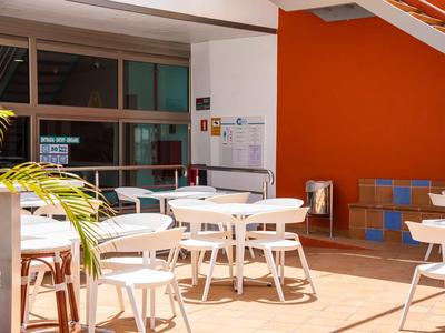 Lichte buitenruimte met witte tafels en stoelen tegen een oranje muur.