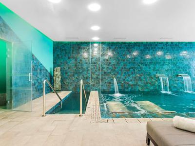 Binnenzwembad met turquoise tegels, waterstralen en ligstoelen in een moderne wellnessruimte.