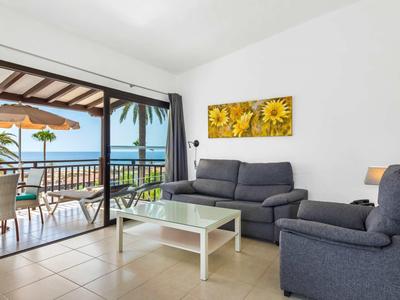 Lichte woonkamer met bank, fauteuil, glazen salontafel en balkon met uitzicht op zee.