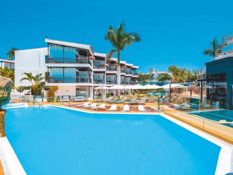 Moderne hotelzwembad met ligstoelen en palmbomen onder een heldere blauwe lucht.